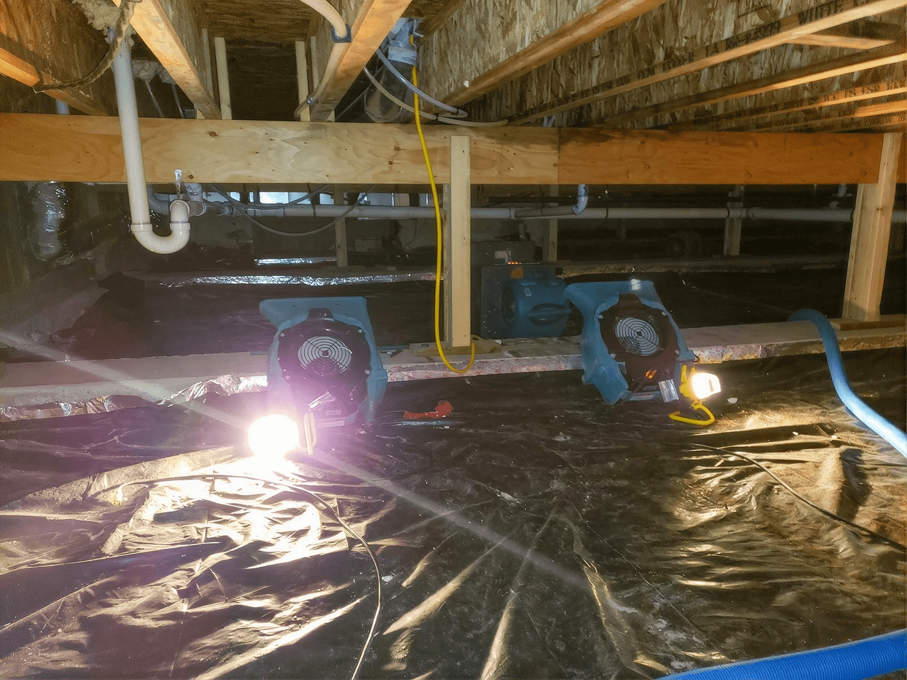 Crawl space equipped with industrial air movers and drying equipment operating over a vapor barrier, with exposed floor joists, plumbing, and ductwork visible as part of a professional moisture mitigation and drying process.